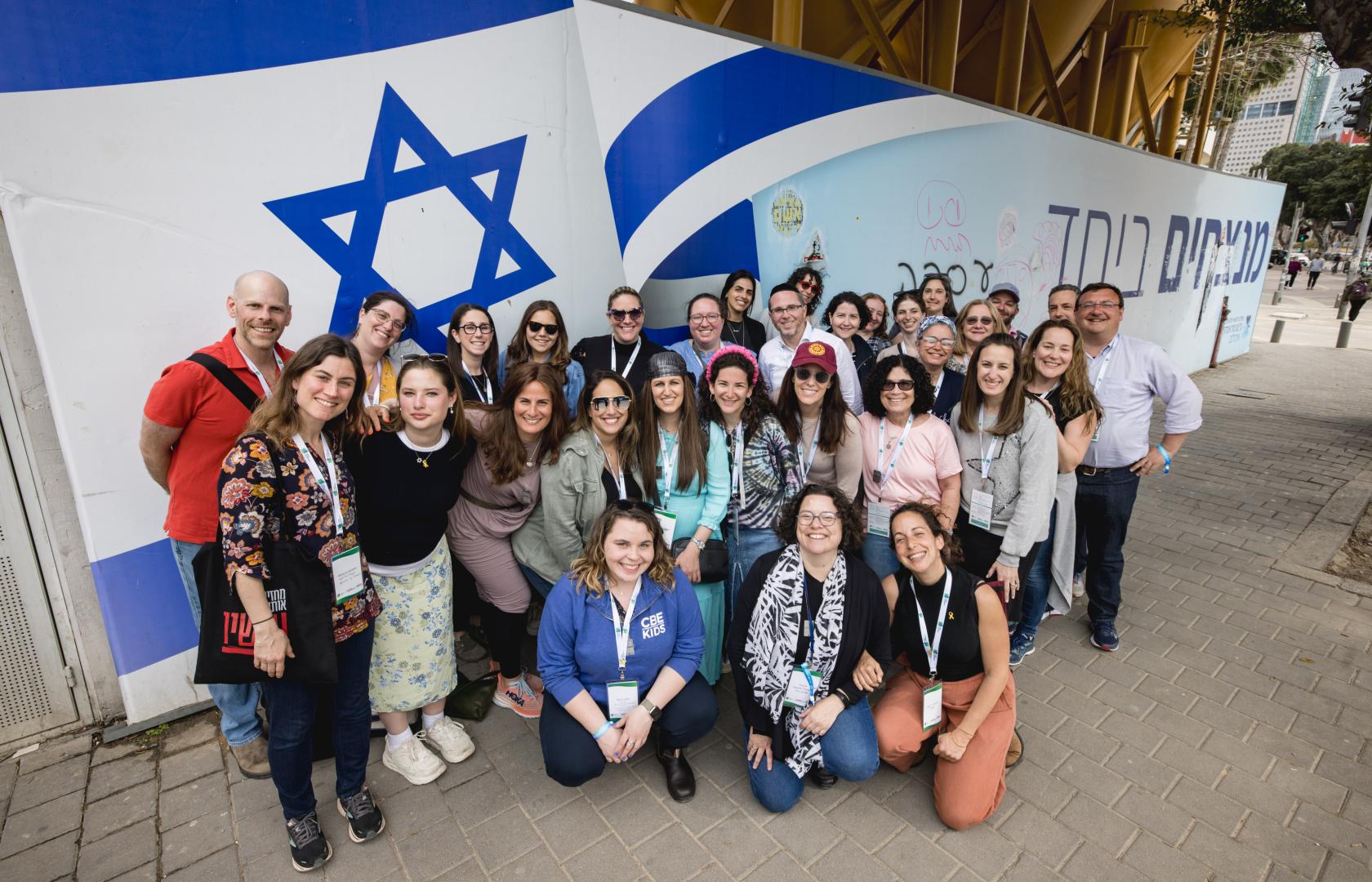 A group photo of educators and staff members smiling in front of a giant Israeli flag