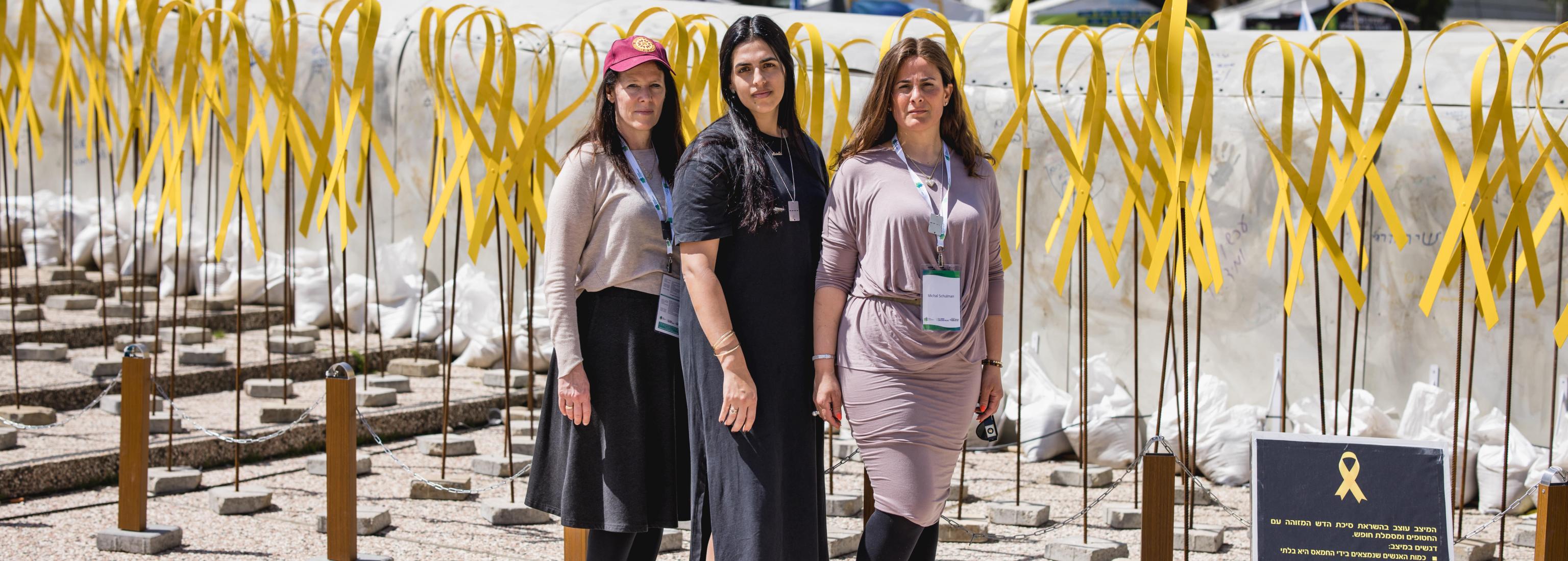 Three female educators stand outside with large yellow ribbons in the background