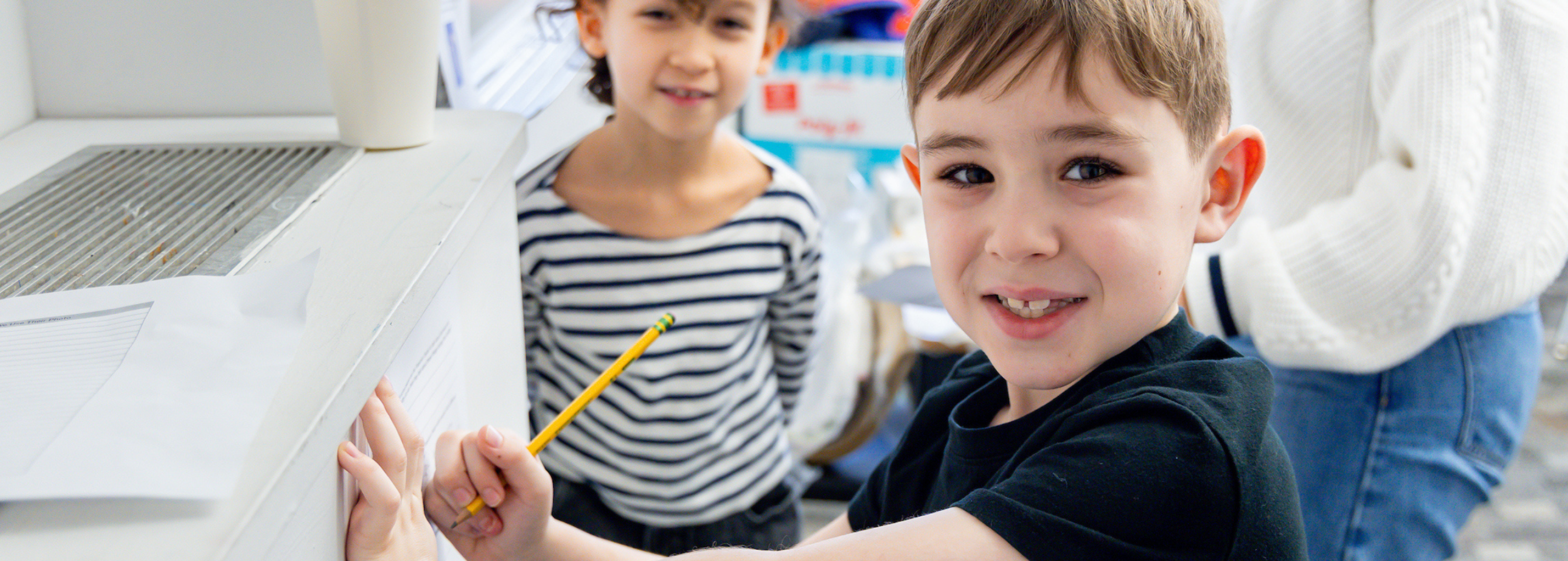 two young children - a boy and a girl - smile at the camera, the boy is in the foreground holding a pencil and paper