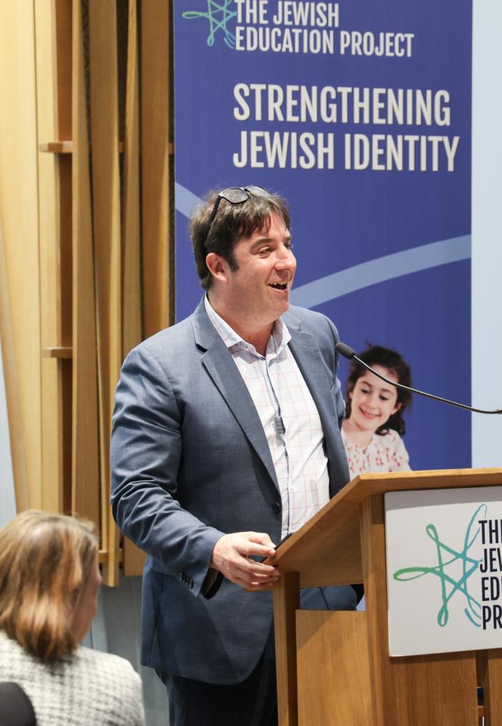 CEO David Bryfman stands in a suit at a podium, smiling, talking to a crowd with The Jewish Education Project logo