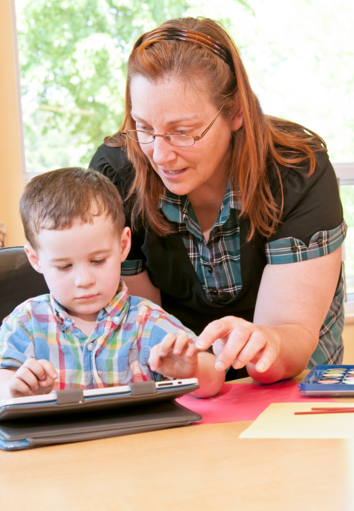 A teacher stands over a young boy and helps him on an ipad