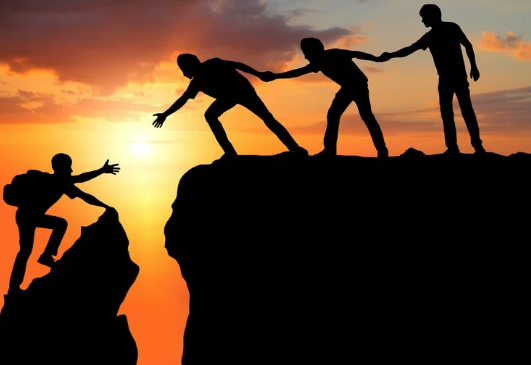 Silhouettes of people helping each other climb up a cliff overlooking a sunset