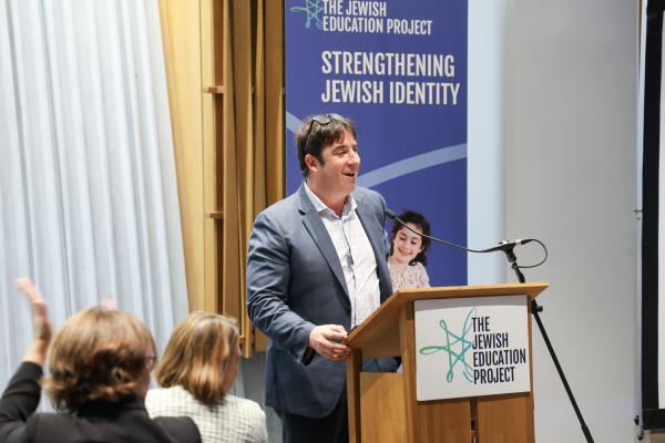 CEO David Bryfman stands in a suit at a podium, smiling, talking to a crowd with The Jewish Education Project logo