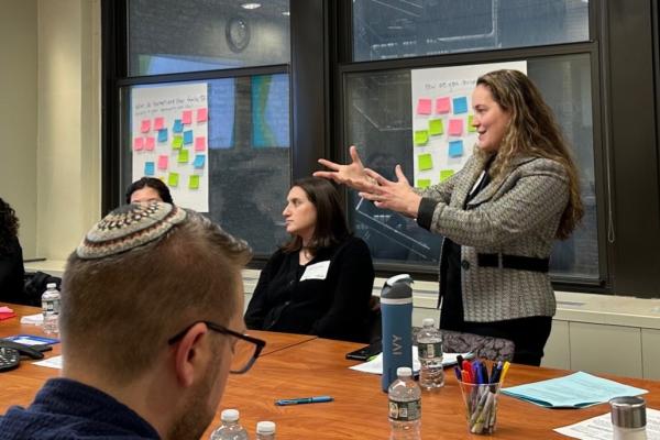 An educator stands and talks in front of a wall of sticky notes with people around a table